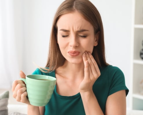 Junge Frau mit Kaffee in den Händen fasst sich an die Wange aufgrund von Zahnspangenschmerzen