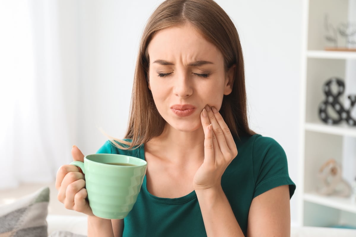 Junge Frau mit Kaffee in den Händen fasst sich an die Wange aufgrund von Zahnspangenschmerzen