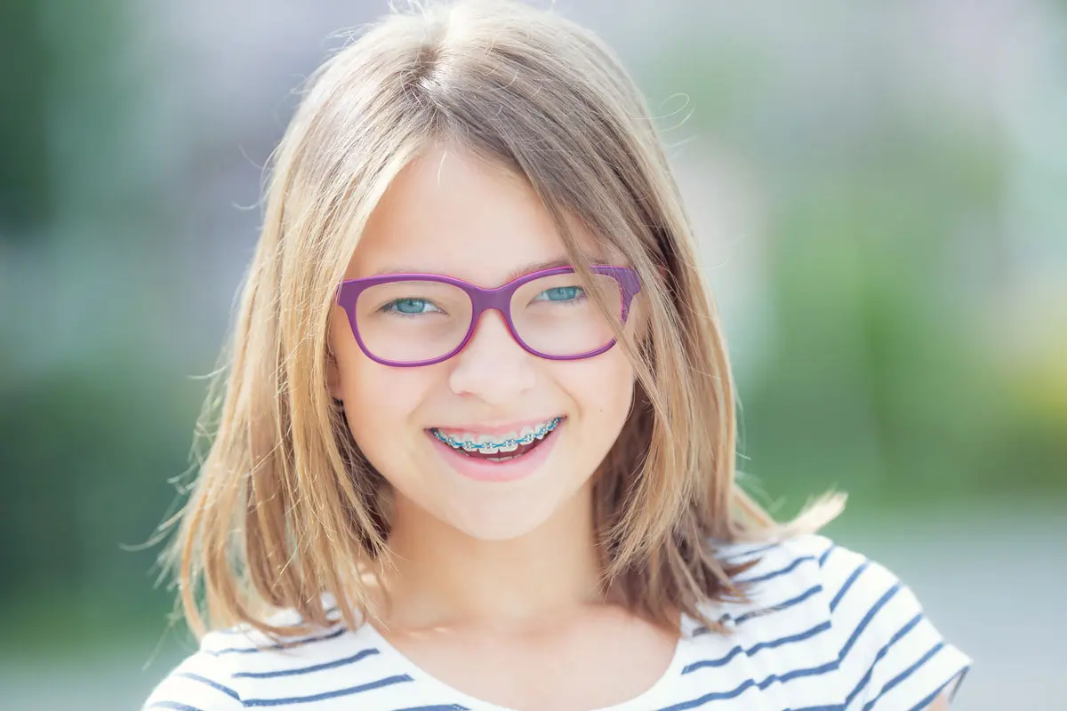 blondes Kind mit lila-farbiger Brille und einer festen Zahnspange mit blauen Gummis